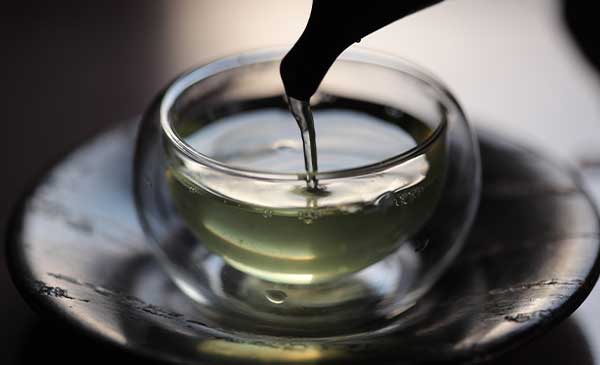 A steaming cup of sencha tea beside fresh green tea leaves.
