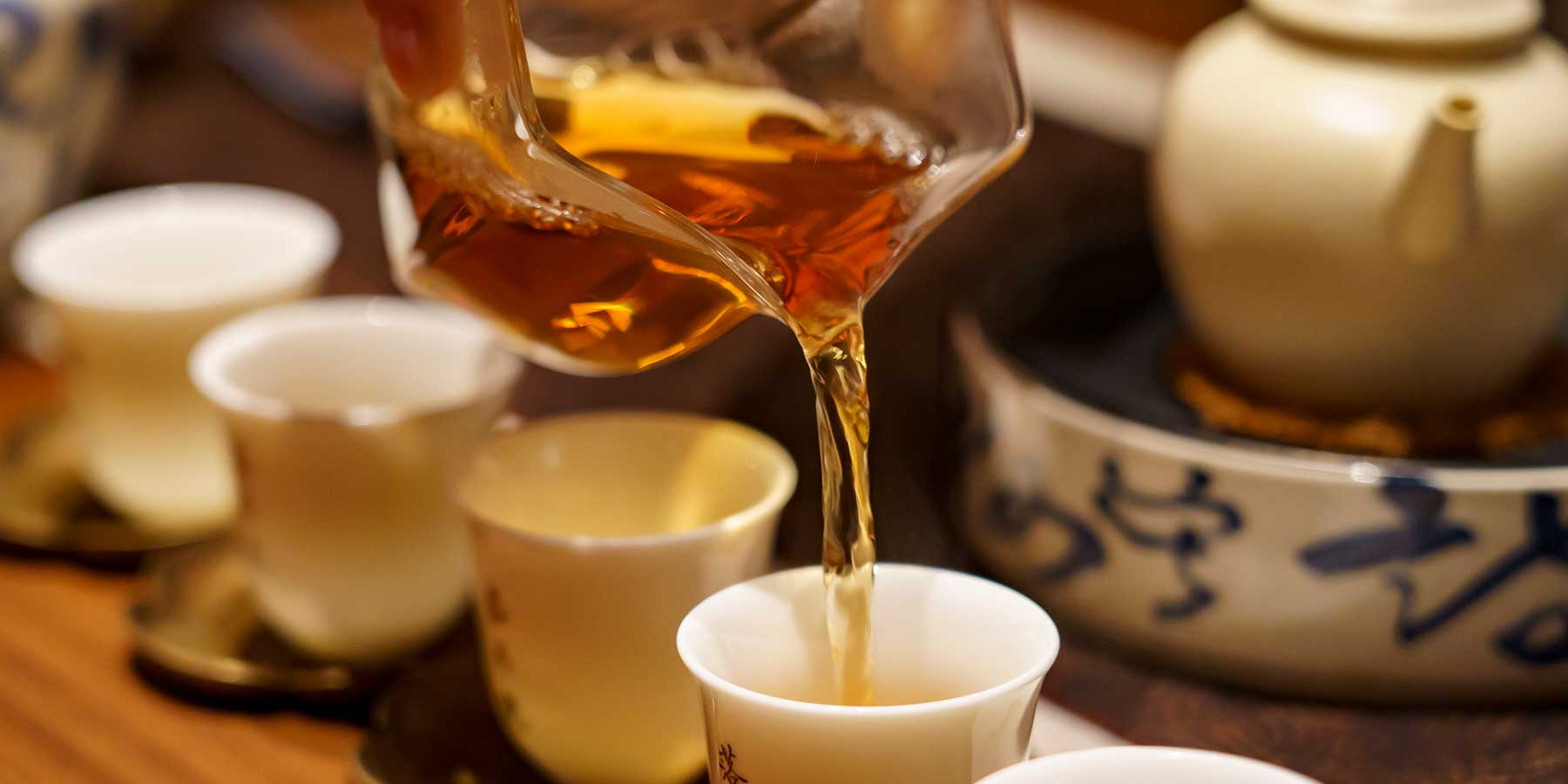 Formosa oolong tea being poured from a teapot into a cup with other cups and a teapot in the background.