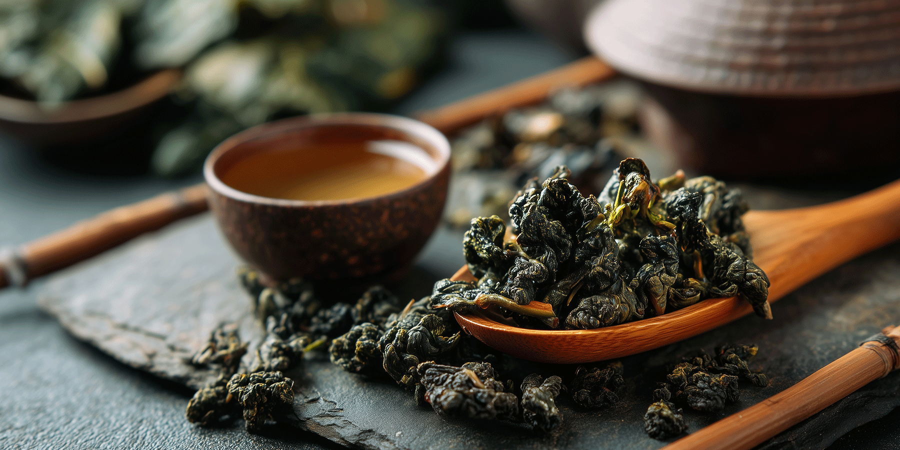 Rolled dark ti kuan yin tea leaves displayed on a wooden spoon and scattered on a slate surface, with a small bowl of brewed tea in the background.